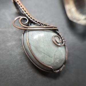 A detailed close-up of Fenrir's Ash, a wire-wrapped pendant with a smooth oval grey serpentine stone encased in intricate copper wire swirls, resting on a dark surface.