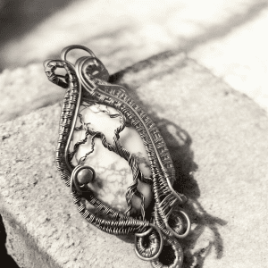 Yggdrasil’s Rest is a handmade pendant with an intricately woven wire forming tree-like shapes and spirals around a white stone with dark veins, set on a rough, light stone—ideal for lovers of unique jewellery.
