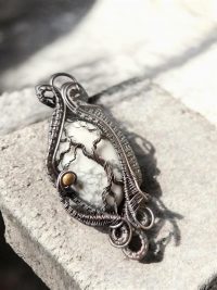 Yggdrasil’s Rest is a handmade pendant with an intricately woven wire forming tree-like shapes and spirals around a white stone with dark veins, set on a rough, light stone—ideal for lovers of unique jewellery.