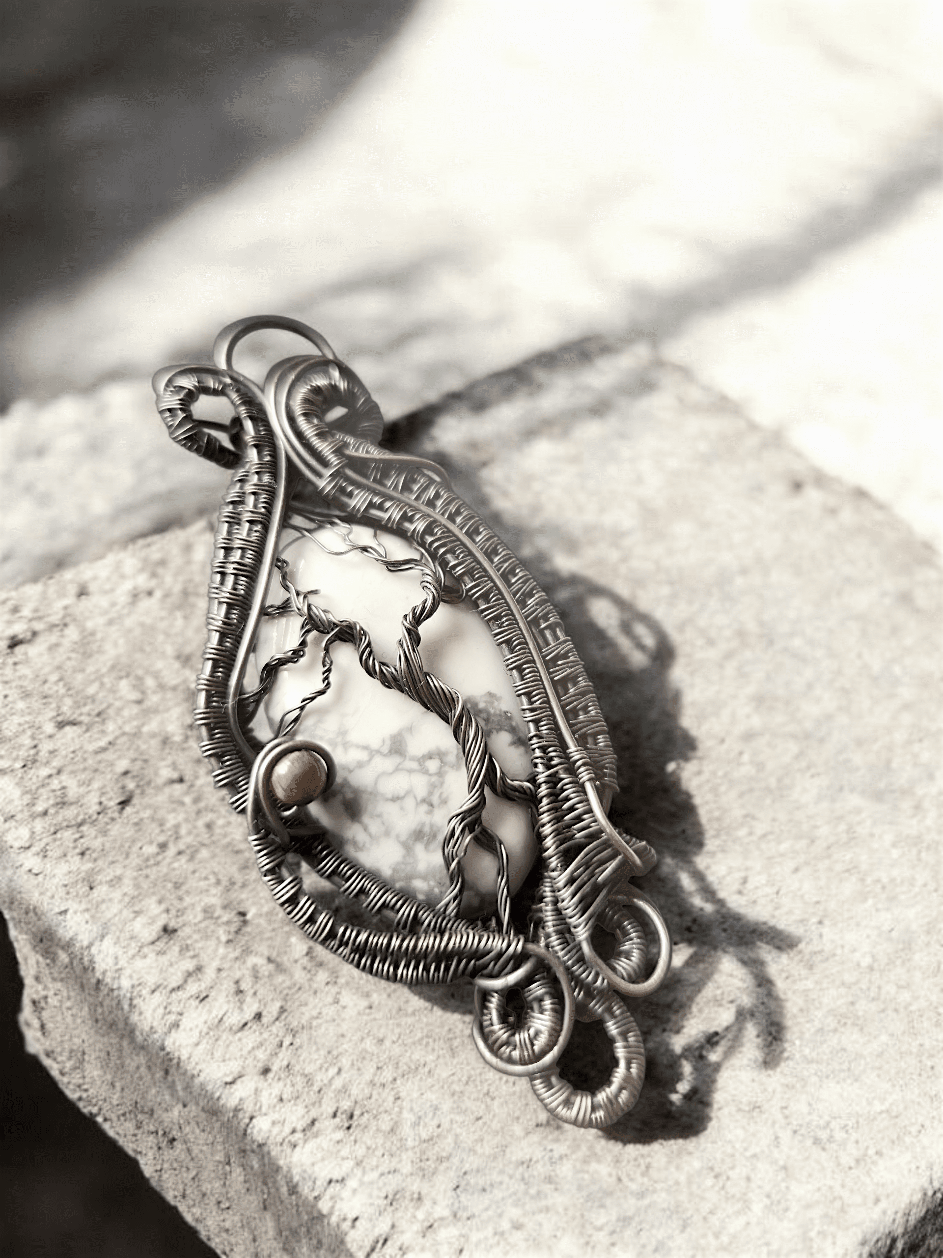 Yggdrasil’s Rest is a handmade pendant with an intricately woven wire forming tree-like shapes and spirals around a white stone with dark veins, set on a rough, light stone—ideal for lovers of unique jewellery.