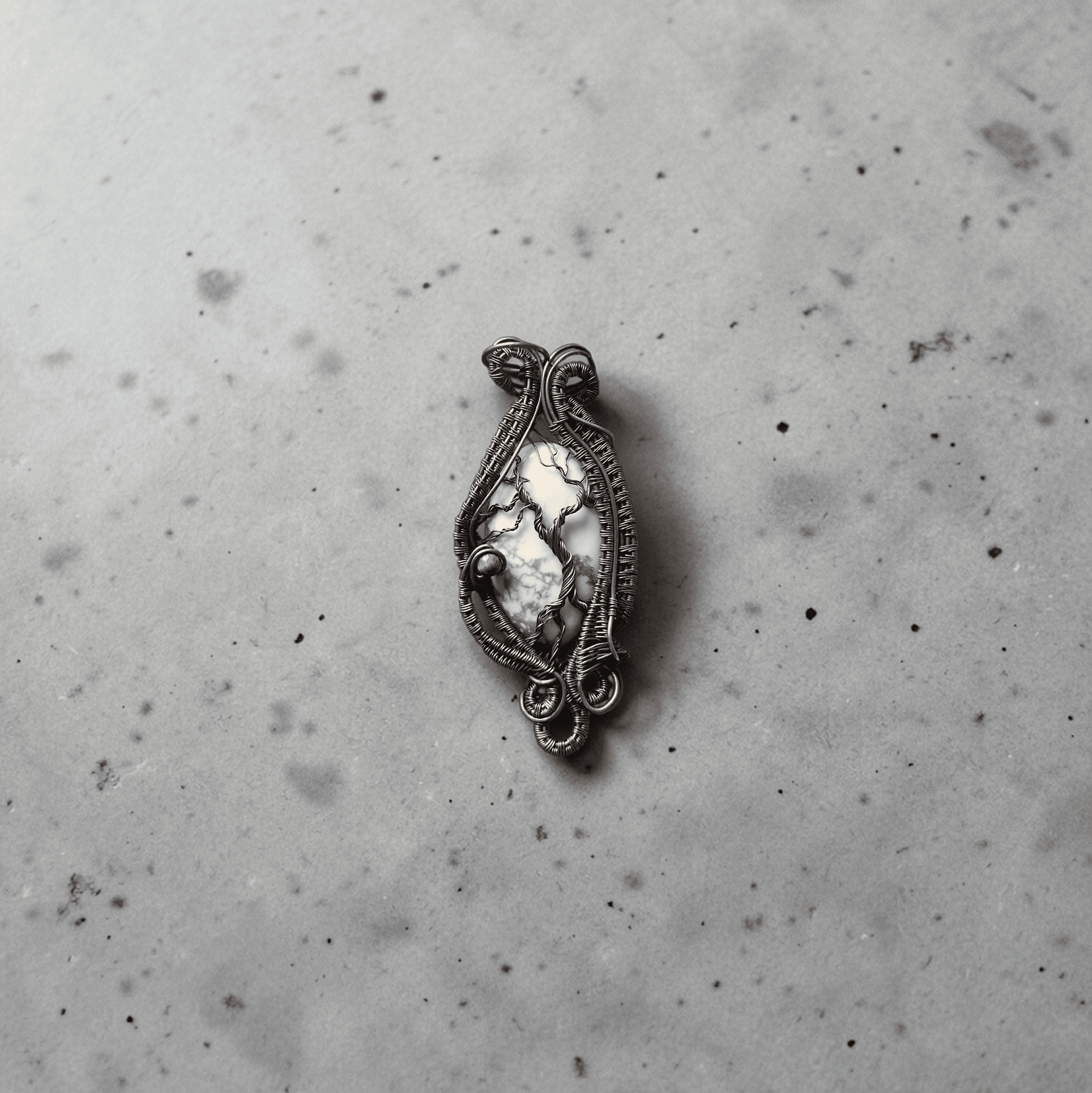Yggdrasil’s Rest, a handmade wire-wrapped pendant with a marbled white and grey stone, rests on a lightly speckled grey surface.