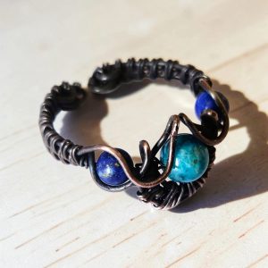 A hand with patterned nails rests on dark fabric, wearing The Wings of Nótt—a silver ring adorned with a blue stone and two smaller blue accents in an ornate, artistic design.