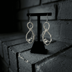 Threads of Urd earrings feature intricate silver wire and bead accents, displayed on a black stand with a textured grey brick background.