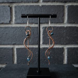 A pair of handmade copper wire wrapped earrings with a curved, swirling design, featuring blue and teal beads, displayed on a black stand in front of a dark brick wall—an elegant piece of jewellery.