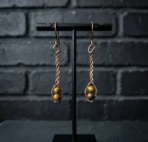Copper wire earrings with twisted stems and brown beads, displayed on a black stand against a white brick wall. Elegant and artistic design.