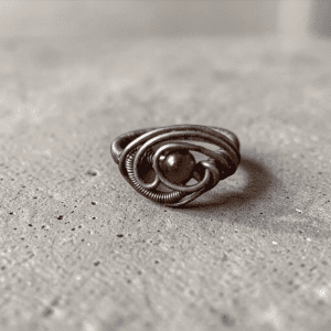 The Seer's Gaze Ring, a handmade wire-wrapped piece with an intricate swirling design and a small brownish stone at its centre, rests on a textured concrete surface.