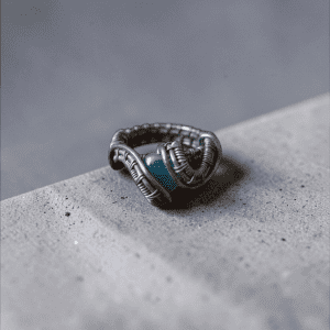 Rán’s Jewel, a handcrafted wire-wrapped ring featuring an intricate design and a striking blue stone centrepiece, rests on a smooth light grey surface with a softly blurred grey background.