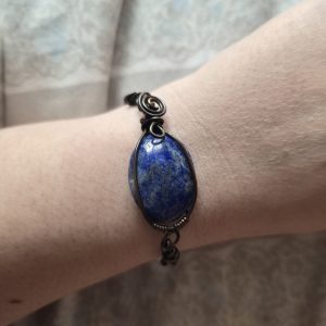 A close-up shows a wrist wearing Fenrir’s Chain, a bracelet featuring a large oval blue gemstone wrapped in dark wire with decorative wirework, set against a pale patterned background.