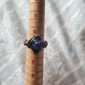 The Vallar-Stjarna silver ring, featuring a blue heart-shaped stone, is shown on a wooden mandrel at approximately size 8. The softly blurred background highlights its elegance.