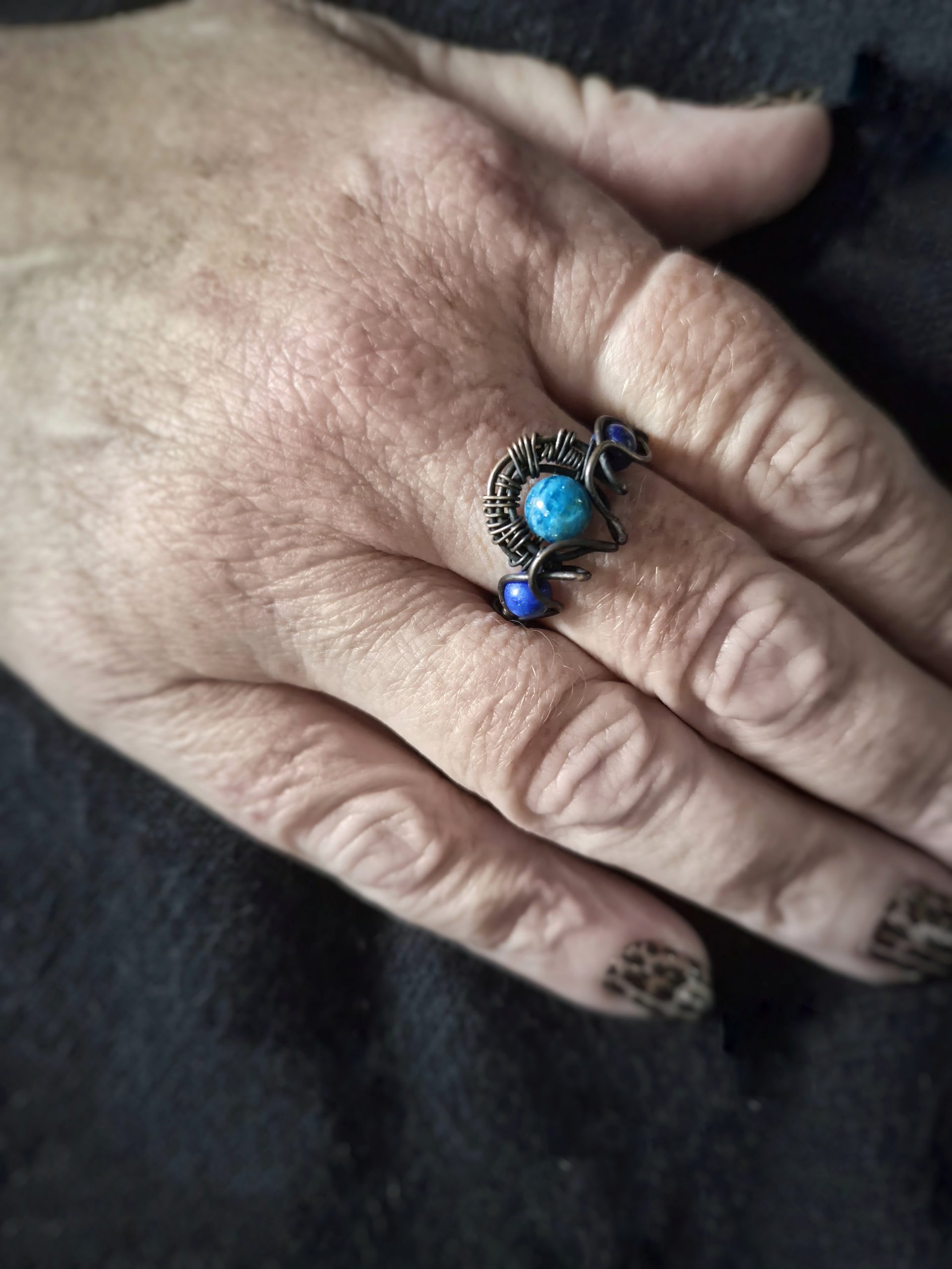 A hand with patterned nails rests on dark fabric, wearing The Wings of Nótt—a silver ring adorned with a blue stone and two smaller blue accents in an ornate, artistic design.