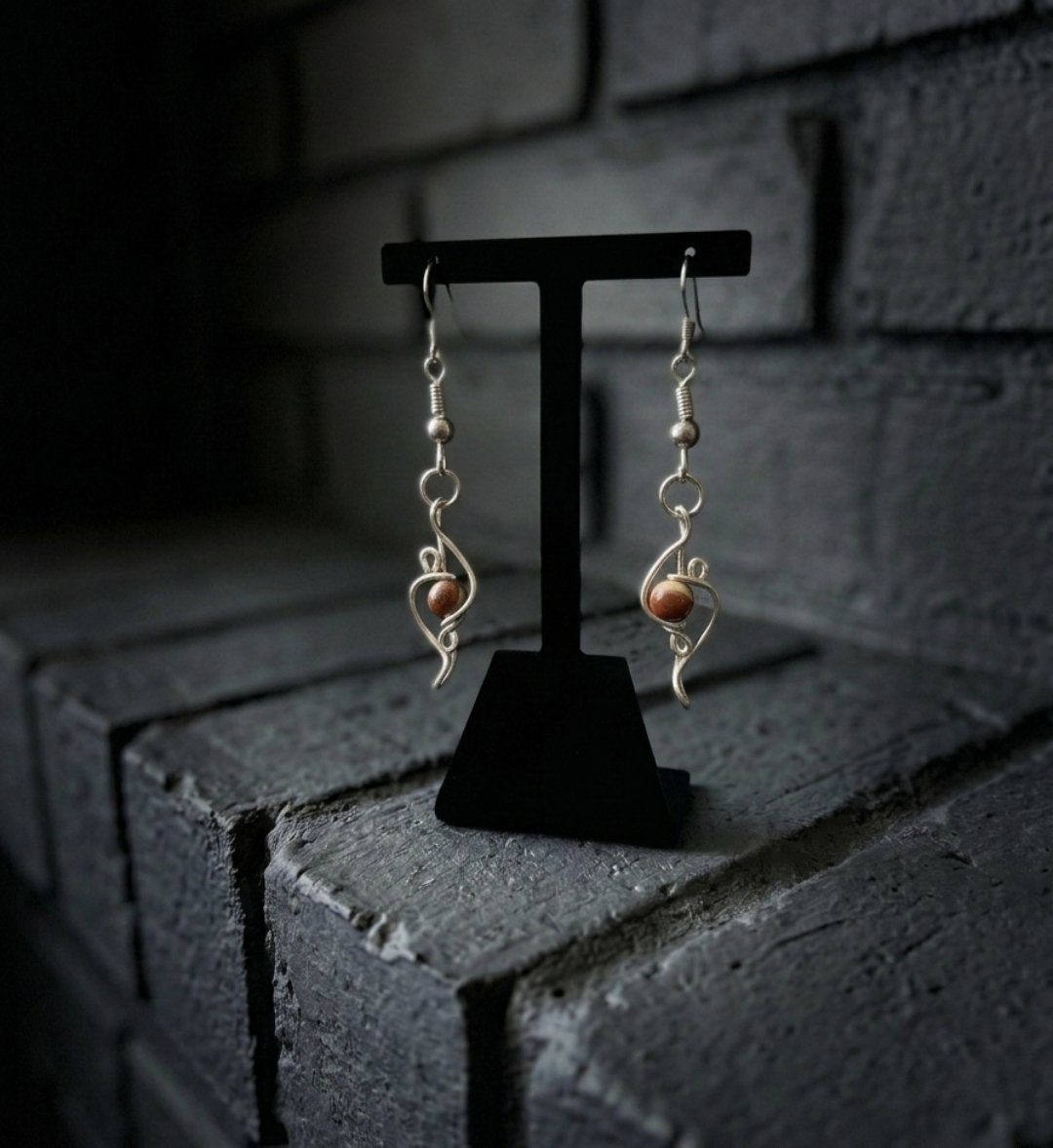 Loki’s Spark: Silver wire earrings with brown beads, showcased on a black stand before grey brick blocks.