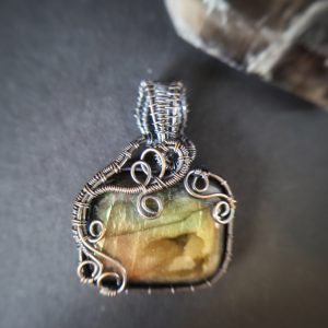 The Shadow Björn: a rectangular gemstone pendant, intricately wrapped in swirling silver wire, displayed on a dark background. A portion of another object appears in the top right corner.
