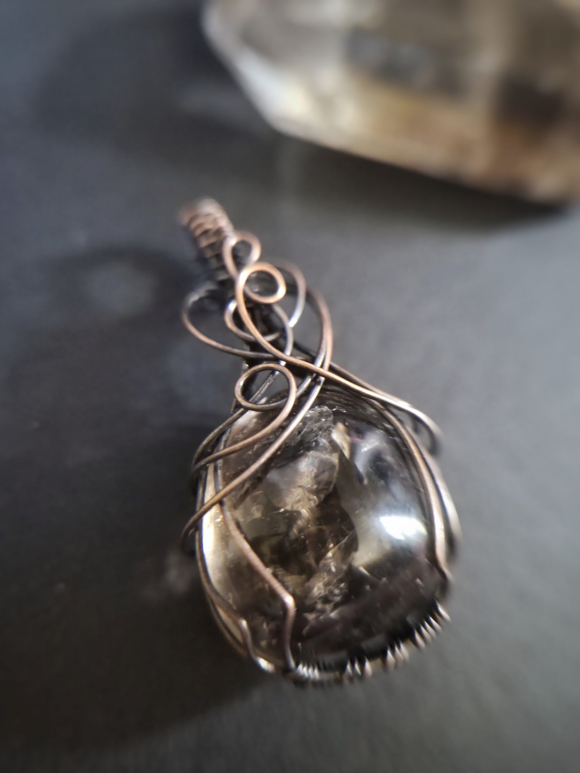 A close-up of Mani's Shadow pendant shows a polished stone wrapped in intricate wire loops, with a blurred background that highlights the jewellery piece.
