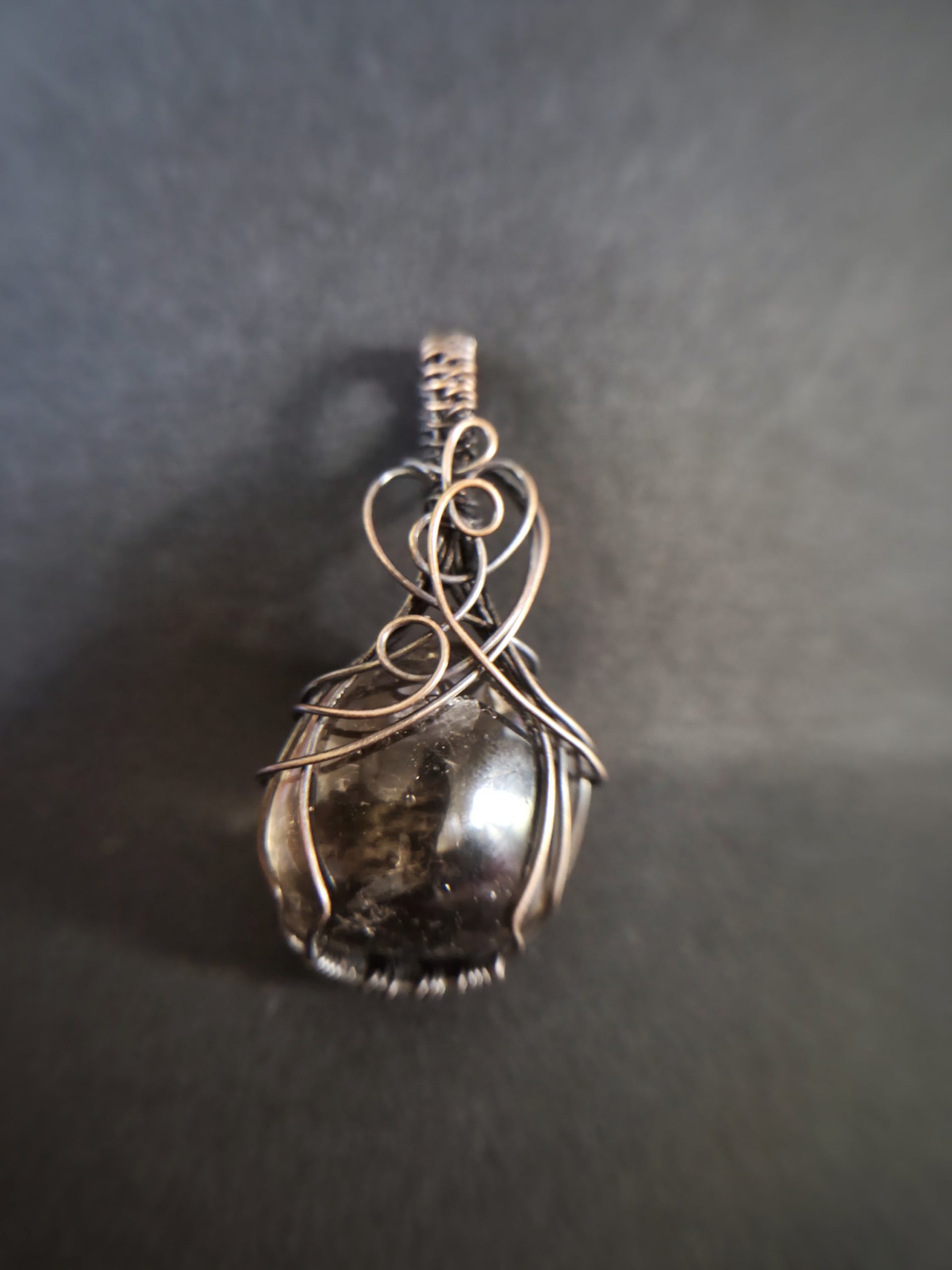 A close-up of Mani's Shadow pendant shows a polished stone wrapped in intricate wire loops, with a blurred background that highlights the jewellery piece.