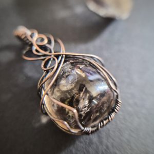 A close-up of Mani's Shadow pendant shows a polished stone wrapped in intricate wire loops, with a blurred background that highlights the jewellery piece.