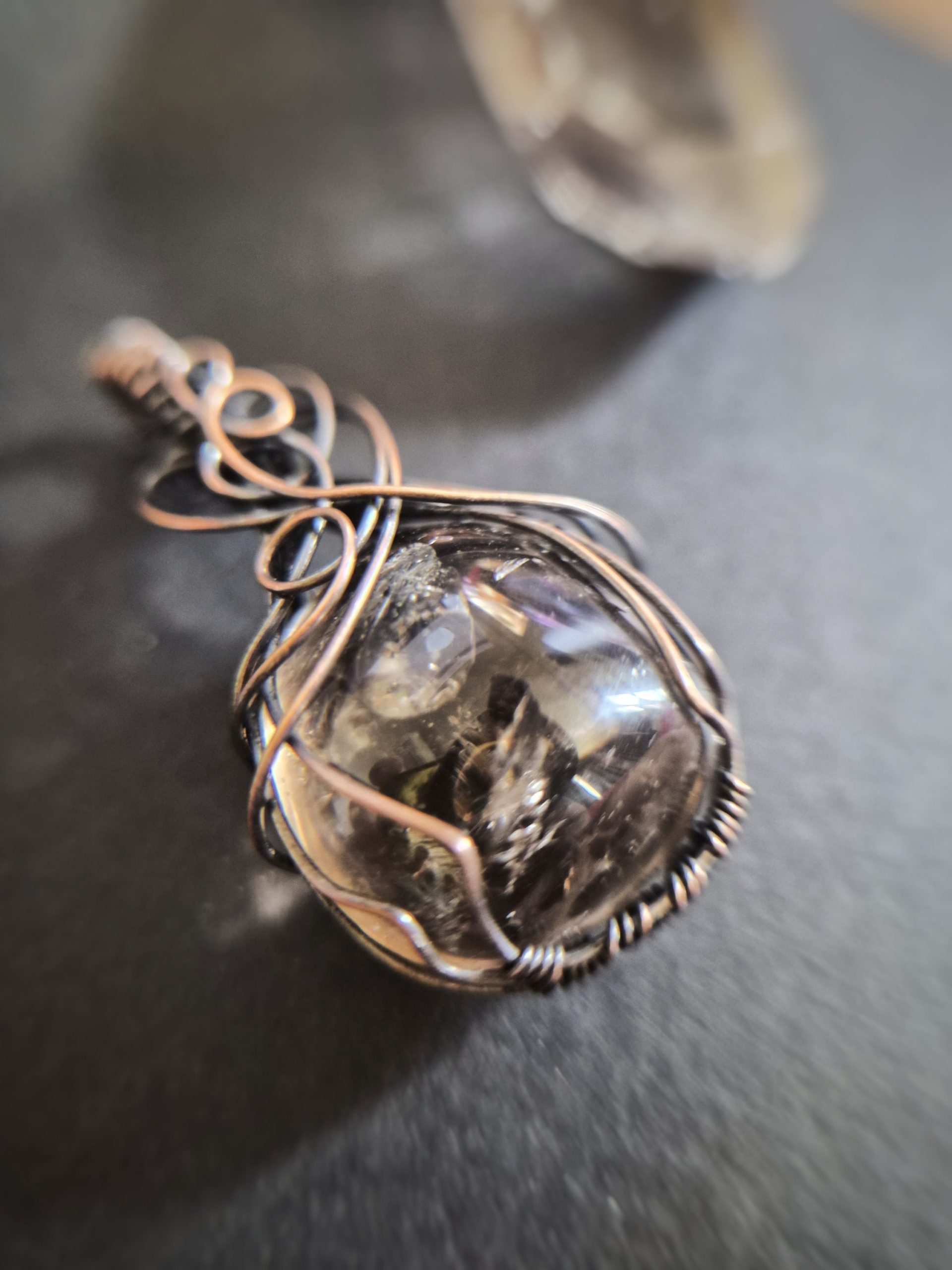 A close-up of Mani's Shadow pendant shows a polished stone wrapped in intricate wire loops, with a blurred background that highlights the jewellery piece.