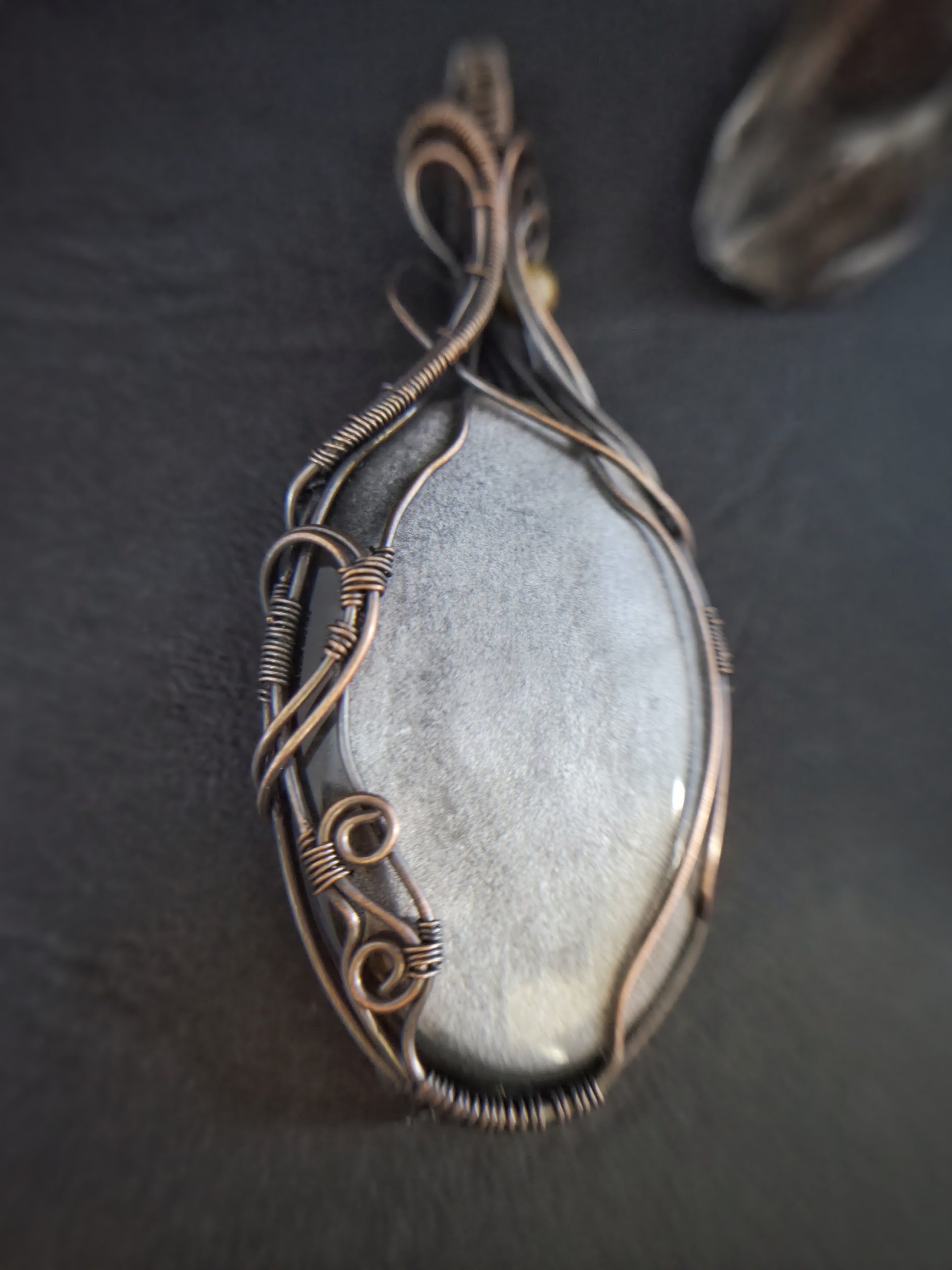 A close-up of Völva's Eye, a smooth oval grey stone pendant wrapped in intricate copper wire, resting on a dark surface. The softly blurred background highlights the elegance of this handcrafted jewellery.