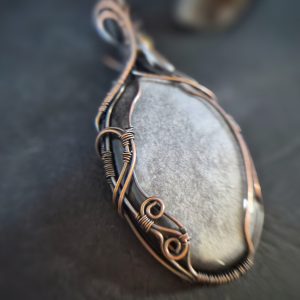 A close-up of Völva's Eye, a smooth silver sheen obsidian stone pendant hand wrapped in intricate copper wire, resting on a dark surface. The softly blurred background highlights the elegance of this handcrafted jewellery.