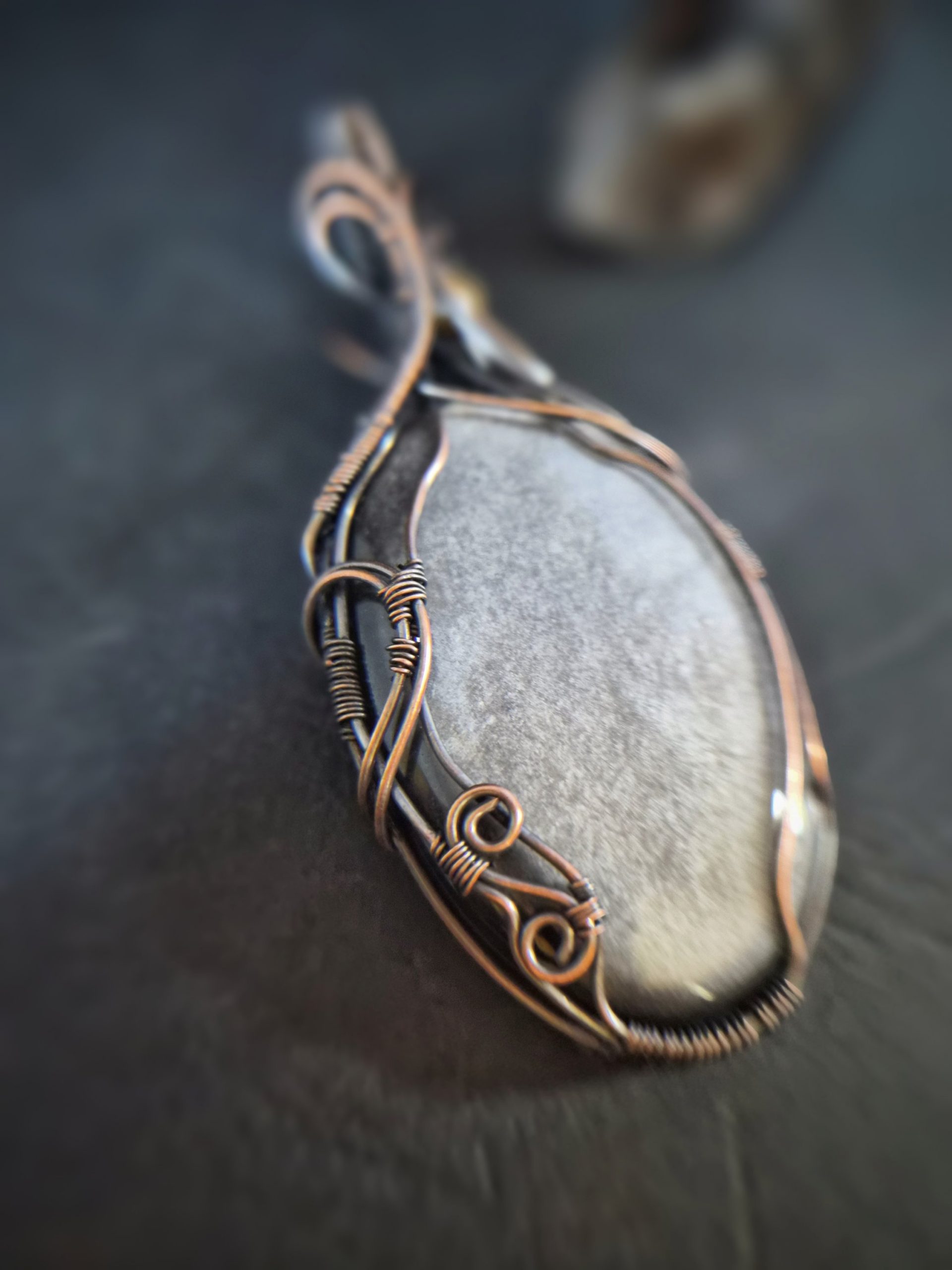A close-up of Völva's Eye, a smooth silver sheen obsidian stone pendant hand wrapped in intricate copper wire, resting on a dark surface. The softly blurred background highlights the elegance of this handcrafted jewellery.
