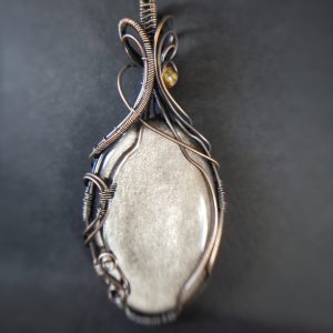 A close-up of Völva's Eye, a smooth oval grey stone pendant wrapped in intricate copper wire, resting on a dark surface. The softly blurred background highlights the elegance of this handcrafted jewellery.