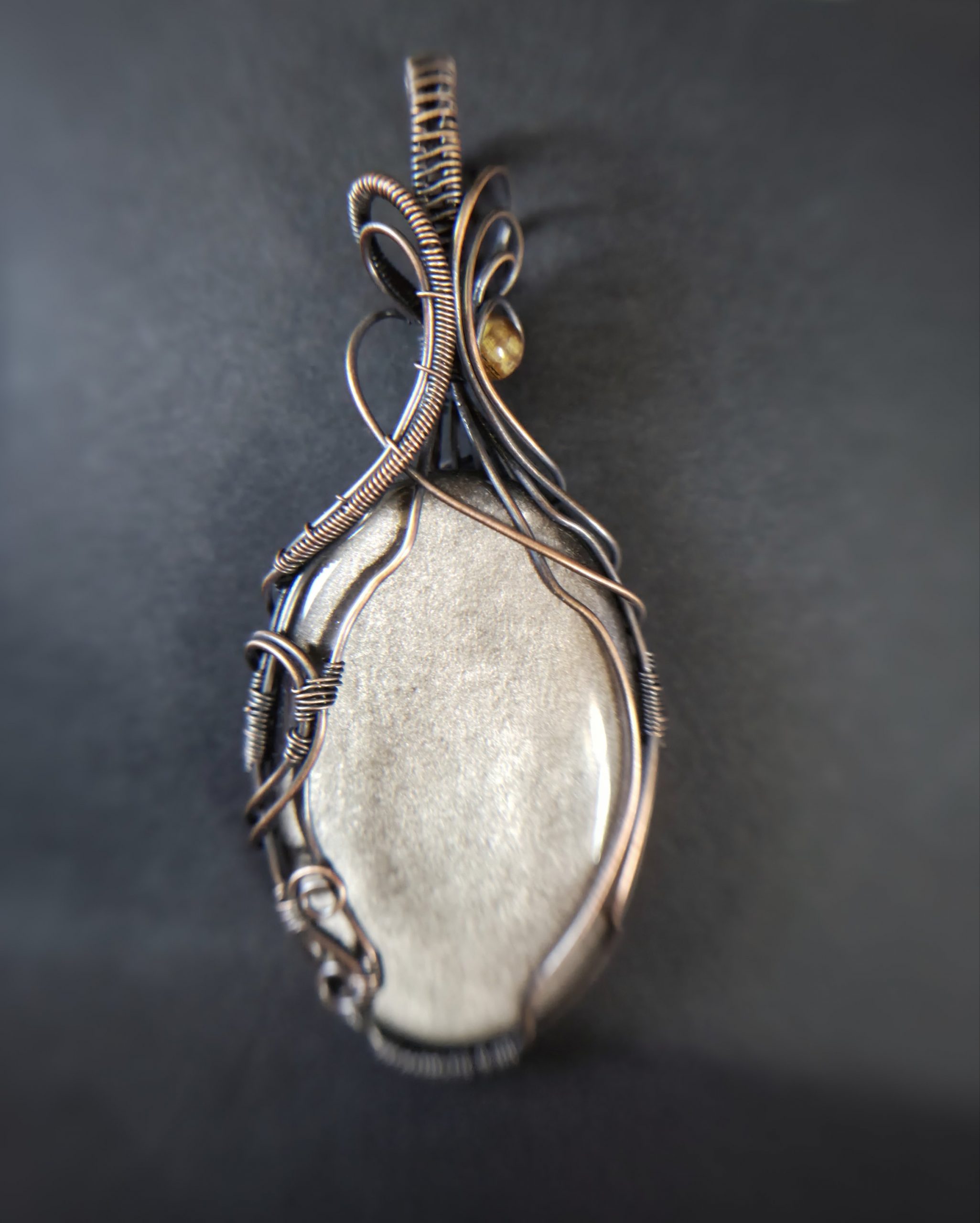 A close-up of Völva's Eye, a smooth oval grey stone pendant wrapped in intricate copper wire, resting on a dark surface. The softly blurred background highlights the elegance of this handcrafted jewellery.