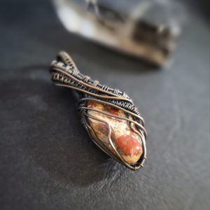 Muspelheim’s Ember: A pendant featuring a polished stone with reddish-orange spots, wrapped in intricate dark wire. Displayed on a dark surface, with a blurred object in the background.