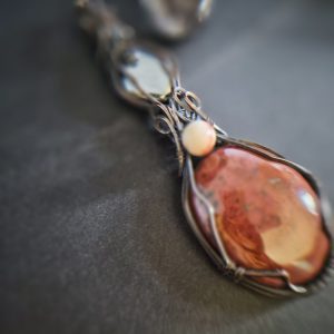 A close-up of Freyja’s Hearthstone, a handmade pendant featuring polished red jasper and haematite, intricately wire wrapped with a small round bead accent, displayed on a dark surface.