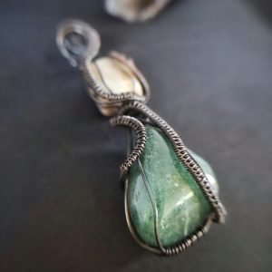 The Seiðr Wand features a green teardrop stone wrapped in intricate wirework and a smaller beige stone above, forming a striking pendant against a dark background.