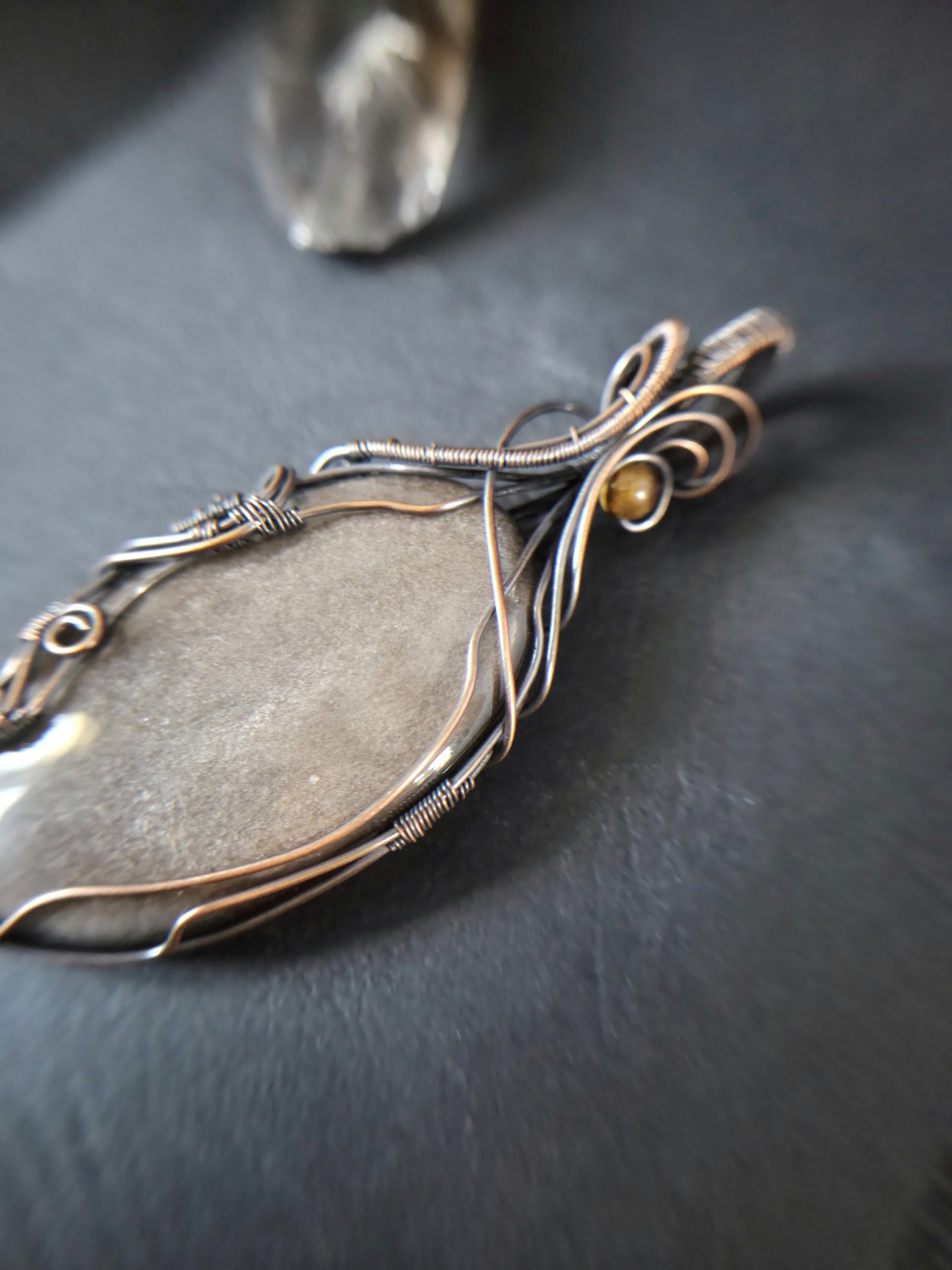 A close-up of Völva's Eye, a smooth oval grey stone pendant wrapped in intricate copper wire, resting on a dark surface. The softly blurred background highlights the elegance of this handcrafted jewellery.