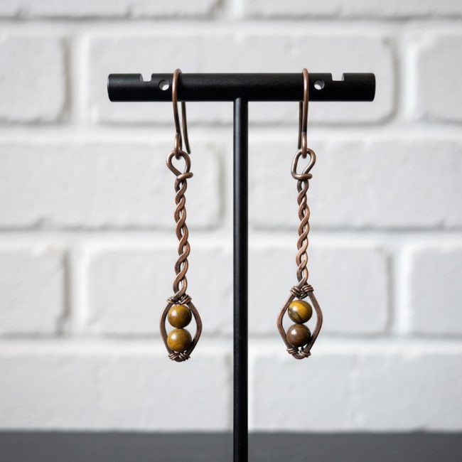 Copper wire earrings with twisted stems and brown beads, displayed on a black stand against a white brick wall. Elegant and artistic design.